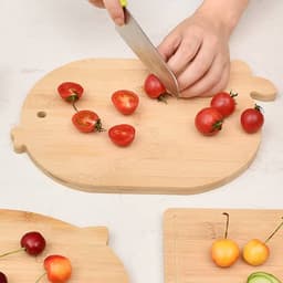 A bamboo cutting board in a playful shape, with cherry tomatoes being chopped and other fruits beside it.
