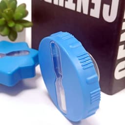 A blue hourglass shower timer with a suction cup, featuring a transparent central section, beside a decorative plant.