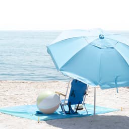 A blue beach umbrella stands beside a beach chair and a brightly coloured beach ball on sand near the water.