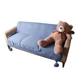 A blue knitted throw blanket with tassels draped over a sofa, alongside a large brown teddy bear.