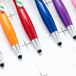 A collection of colourful ballpoint pens in orange, red, blue, purple, and silver, featuring stylus tips and branding.