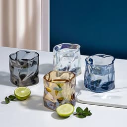 A set of four colourful irregular whiskey cups in blue, clear, amber, and dark blue, accompanied by lime halves and mint leaves.