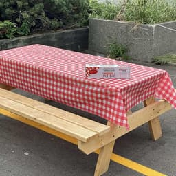 A red and white checkered PEVA tablecloth drapes over a wooden picnic table. A branded box is visible beside it.