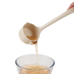 A wheat straw oil filter spoon in a light beige colour pouring liquid into a glass bowl. It features a logo.