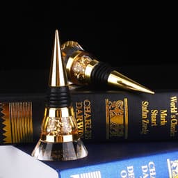 A drink bottle stopper in gold and clear crystal, featuring a sleek design and a black rubber base, rests on a stacked book.