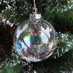 A transparent glass ornament ball with rainbow reflections, hanging from a pine tree branch.