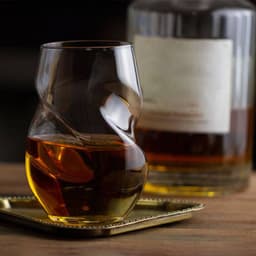 A stemless wine glass with finger indentations, holding amber liquid, sits on a tray beside a bottle.