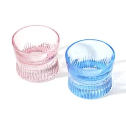 Two crystal whiskey corset glasses, one in pink and one in blue, featuring ribbed designs. Both have a logo.
