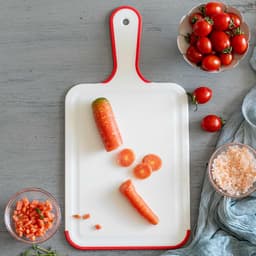 A non-slip plastic cutting board in white with red edges, featuring cut vegetables and cherry tomatoes on a wooden surface.