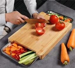 A bamboo chopping board featuring two stainless steel trays, with sliced vegetables and tomatoes on the surface.