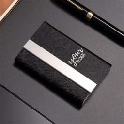 A black PU leather business card case with a silver stripe and a logo, resting on a dark surface beside a pen.