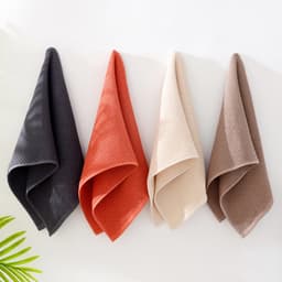 Four face towels in dark grey, terracotta, cream, and light brown, hanging on a wall with a plant partially visible.