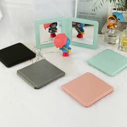A set of square travel makeup mirrors in black, grey, pink, and mint green, arranged with a decorative figure.