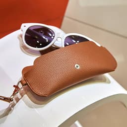 A brown portable glasses pouch with strap beside stylish white sunglasses on a display surface.
