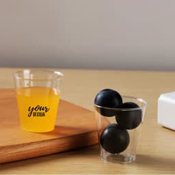 A 2 oz. clear plastic party cup holding yellow liquid next to another cup containing black balls, on a wooden surface.