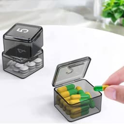 A stack of transparent black pill organiser boxes containing yellow and green capsules, with one box open and a hand holding a capsule.