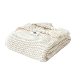A knitted cream-coloured throw blanket featuring decorative tassels and a logo. Soft and textured design.