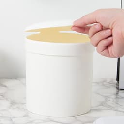 A white mini desktop trash can with a curved lid, placed on a marble surface.
