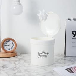 A white mini desktop trash can with a lid, placed on a marble surface, alongside a wooden clock and documents.