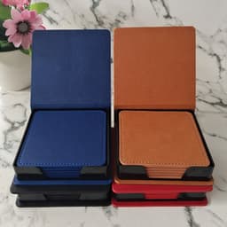 A stack of four PU leather coasters in blue, tan, red, and black, displayed in a holder. A plant is in the background.