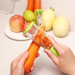 A green peeler with a clear storage box, being used on a carrot, with apples and melons in the background.