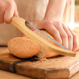 A serrated bread knife with a wooden handle slicing through a sesame seed roll on a wooden cutting board.