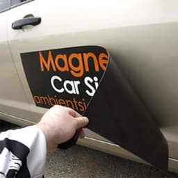A hand holding a custom car magnet, showcasing a black backing and vibrant orange text, ready to be applied to a vehicle.