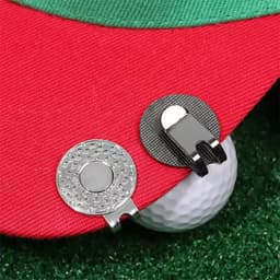 A red golf hat featuring two metal clips, one round and one flat, positioned near a golf ball on green turf.