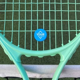 A mint green tennis racket with a personalised blue dampener nestled in its strings.