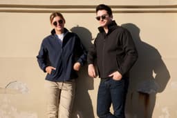 A navy ladies' soft shell jacket and a black men's soft shell jacket are worn by two models against a wall.