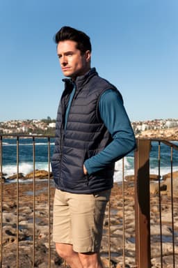 A man stands on rocky beachwearing a navy blue puffer vest and a teal long-sleeve top, with coastline in the background.