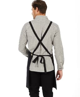 A black bib apron with crossed straps is displayed on a person wearing a checked shirt. The apron features a logo.