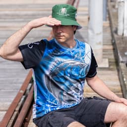 A man wearing a blue and black polyester fishing tee and a green bucket hat, sitting on a dock.