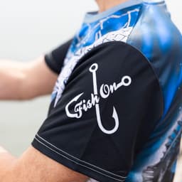 A men's fishing tee in blue and black with a fishing hook design on the sleeve and logo branding.