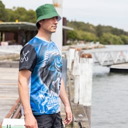 A man wearing a blue and black polyester sublimated fishing tee and a green bucket hat by the water.