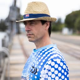 A man wearing a straw hat and a blue patterned long sleeve fishing tee stands by a waterway with a serene expression.