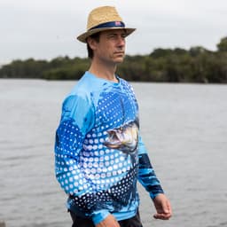 A man wearing a long sleeve blue polyester fishing tee, featuring a fish graphic, near a body of water.