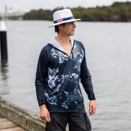 A man stands by the water wearing a navy blue and grey patterned 1/4 zip fishing hoodie and a white hat.