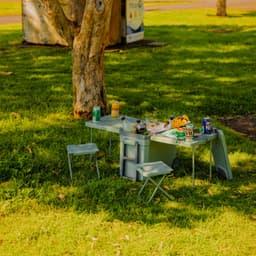 A green cooler box with an attached table and two chairs, set on grass with drinks and snacks around it.