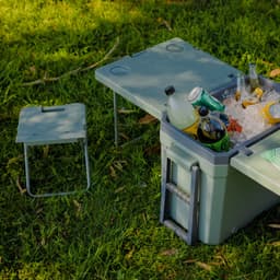 A green cooler box with a table and a small chair, filled with beverages and ice, set on grass.