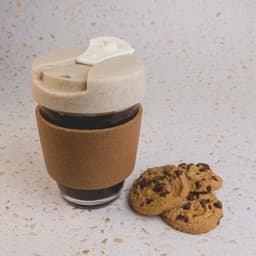 A reusable coffee cup with a cork band and a light-coloured lid, alongside chocolate chip cookies on a countertop.