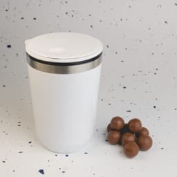 A reusable white coffee cup with a stainless steel band, beside a small pile of brown chocolate balls.