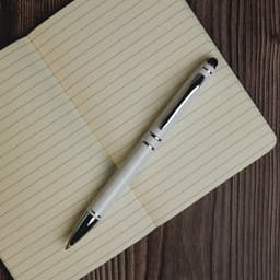 A metal pen in white and black rests atop an open lined notebook on a wooden surface. The pen features a logo.