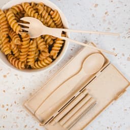 A bamboo cutlery set in a case, featuring a spoon, fork, and chopsticks, alongside a bowl of pasta.