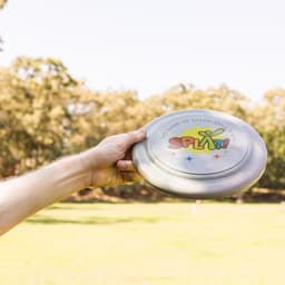 A hand holds a silver frisbee with a colorful logo, set against an outdoor grassy background.