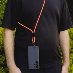 A phone lanyard in bright orange, worn around the neck, holding a smartphone against a green outdoor background.