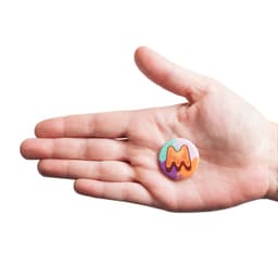 A hand holding a 32mm round badge with vibrant orange, green, and purple colours, featuring a logo.