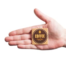 A hand holding a square coffee packet with a brown and gold design, featuring a coffee bean logo.