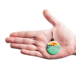 A hand holds a custom printed 38mm round zipper pull in green, featuring a logo and vibrant design.