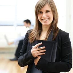A young woman in a black blazer holds a dark padfolio and a pen, smiling in a bright office setting.